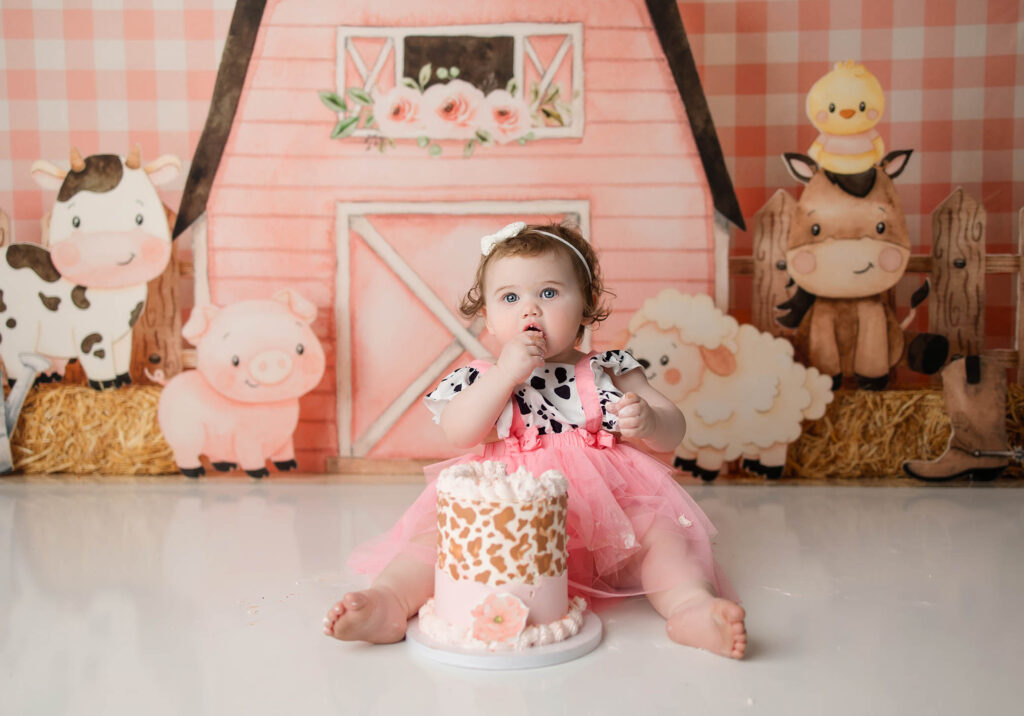 A baby in a pink and cow-print outfit sits on the floor in front of a barnyard-themed backdrop, enjoying a pink and white birthday cake decorated with cow spots. Captured by a talented phoenix maternity photographer, farm animal illustrations complete the scene.