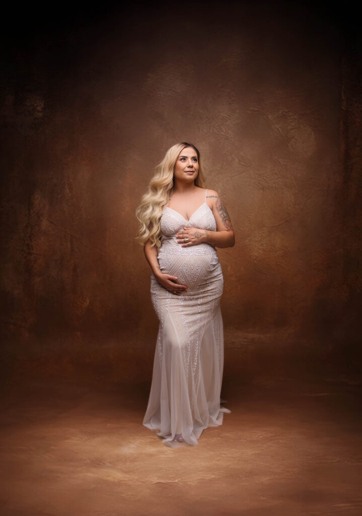 A pregnant woman with long blonde hair stands in a fitted, sleeveless, light-colored gown, holding her belly and gazing slightly upward against a brown textured studio background, captured by a skilled Peoria maternity photographer.