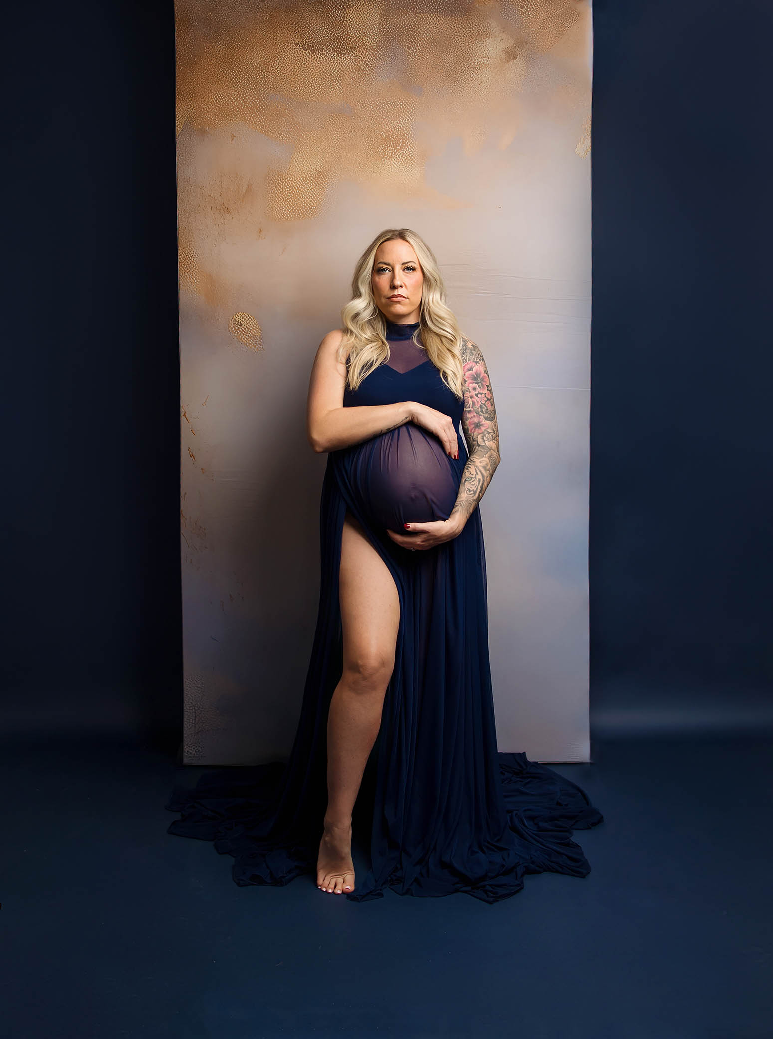 Pregnant woman in sheer navy dress stands barefoot, holding belly against a dark background.