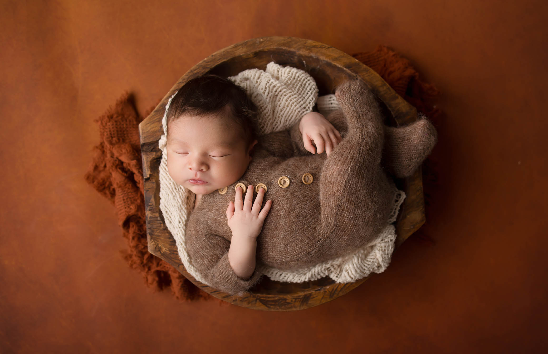 A newborn baby, dressed in a brown knitted onesie, sleeps peacefully curled up in a round wooden bowl lined with soft white and brown blankets on a warm brown background, beautifully captured by a skilled Phoenix newborn photographer.