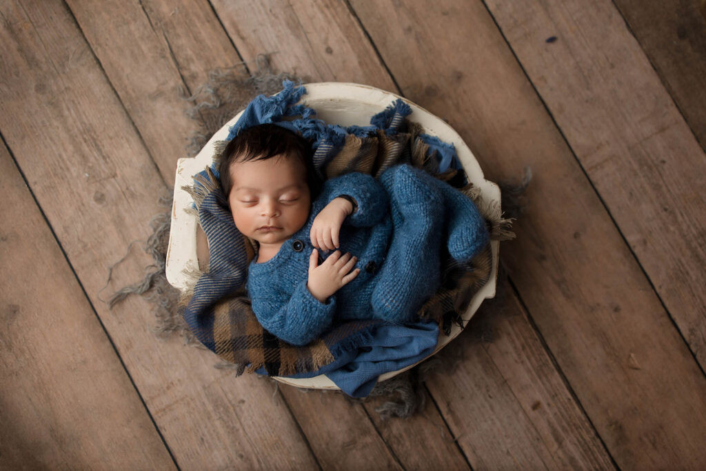 A sleeping baby in a blue outfit lies curled up on a plaid blanket inside a round white bowl, placed on a wooden floor—captured by a talented family photographer Phoenix AZ.