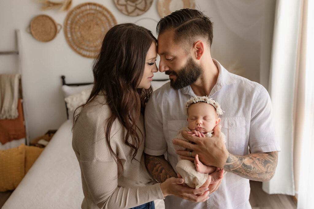 A couple stands close together, gently touching foreheads, as the man cradles a sleeping baby in pink. This warm, intimate scene with soft natural lighting captures the loving bond cherished by any family photographer Phoenix AZ.