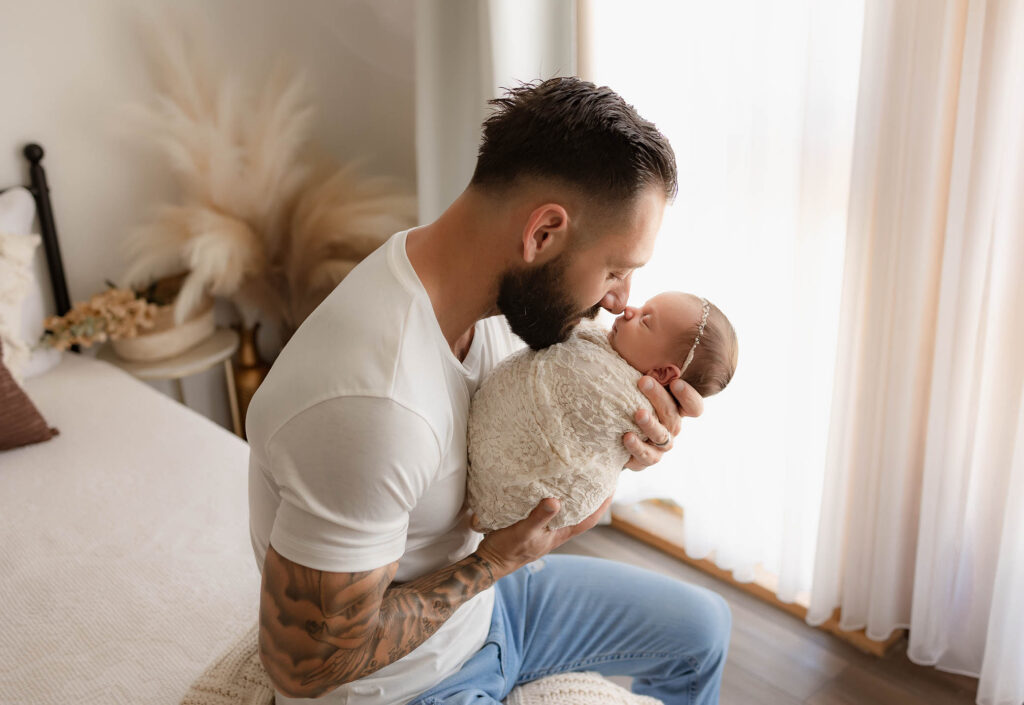A bearded man in a white t-shirt cradles a swaddled newborn, touching noses and smiling, while sitting on the bed of a softly lit room—captured by a talented Phoenix newborn photographer.