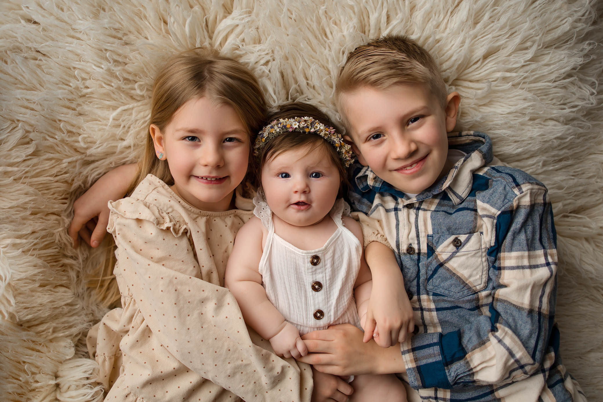 Three young children lie close together on a fluffy, cream-colored rug. Captured by a talented Phoenix newborn photographer, their smiles and relaxed poses highlight the joy and warmth of this special moment.