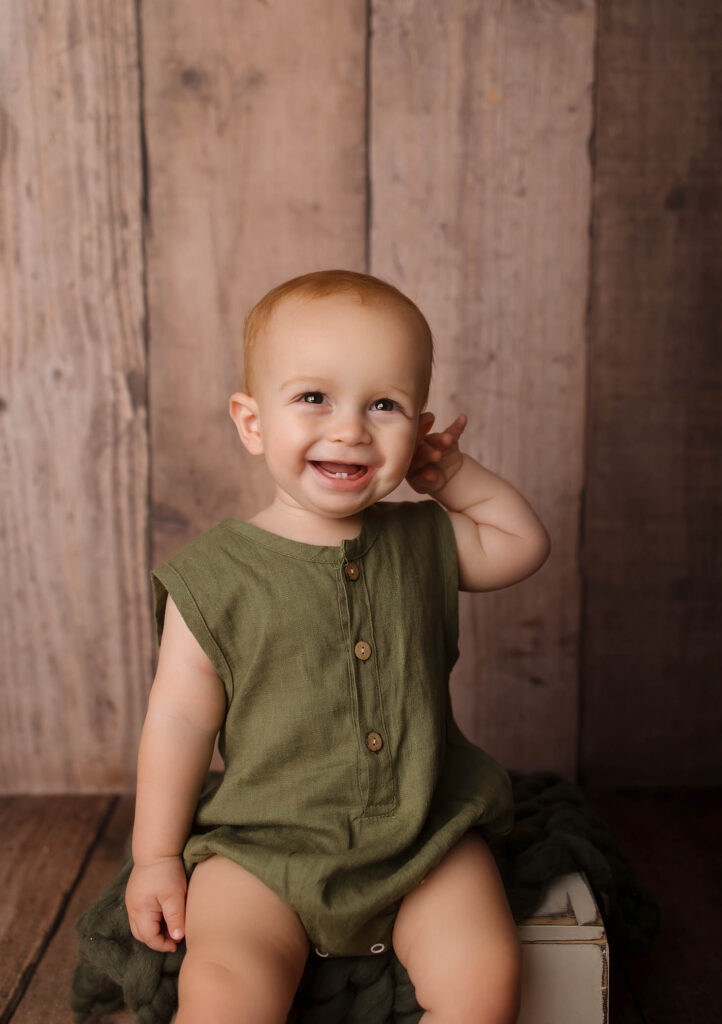 A smiling baby with light skin and short light brown hair wears an olive green romper and sits on a dark green blanket against a rustic wooden background—captured perfectly by a talented family photographer in Phoenix, AZ. One hand is raised near their ear.
