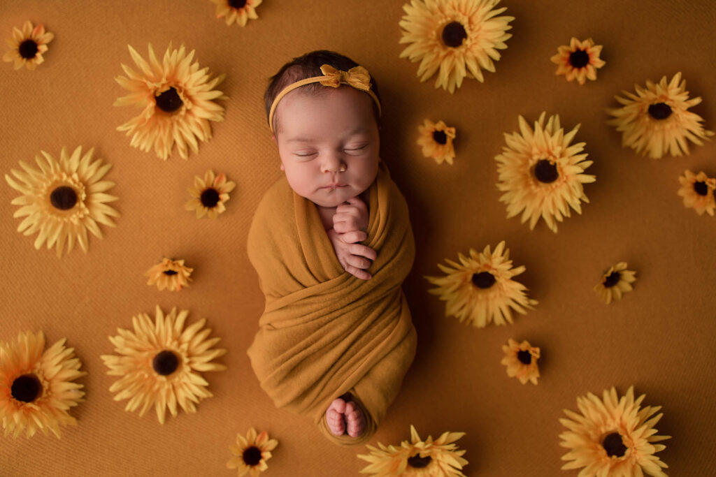A newborn baby wrapped in a mustard-yellow blanket lies on a matching background, surrounded by artificial yellow sunflowers. Captured by a Phoenix newborn photographer, the baby wears a yellow bow headband and sleeps peacefully with eyes closed.
