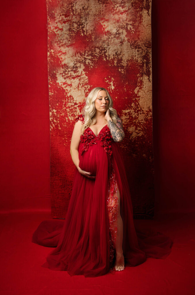 A pregnant woman with long blonde hair in a flowing red gown stands barefoot against a textured red and gold backdrop, gently cradling her belly and touching her hair, photographed by a talented phoenix maternity photographer. Visible tattoos grace her left arm.