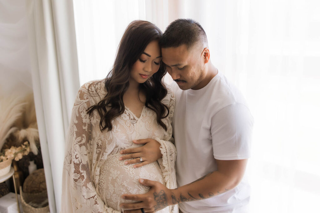A couple stands by a bright window, both gently cradling the woman's pregnant belly. Captured by a Scottsdale maternity photographer, she wears a lace dress and has long hair, while he rests his hand lovingly on her bump.