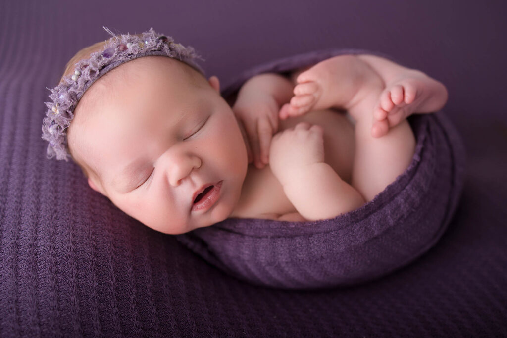 A sleeping newborn baby with a purple headband is curled up on a textured purple blanket, partially wrapped in matching purple fabric. Captured by a phoenix newborn photographer, the baby’s arms and legs are gently tucked in.