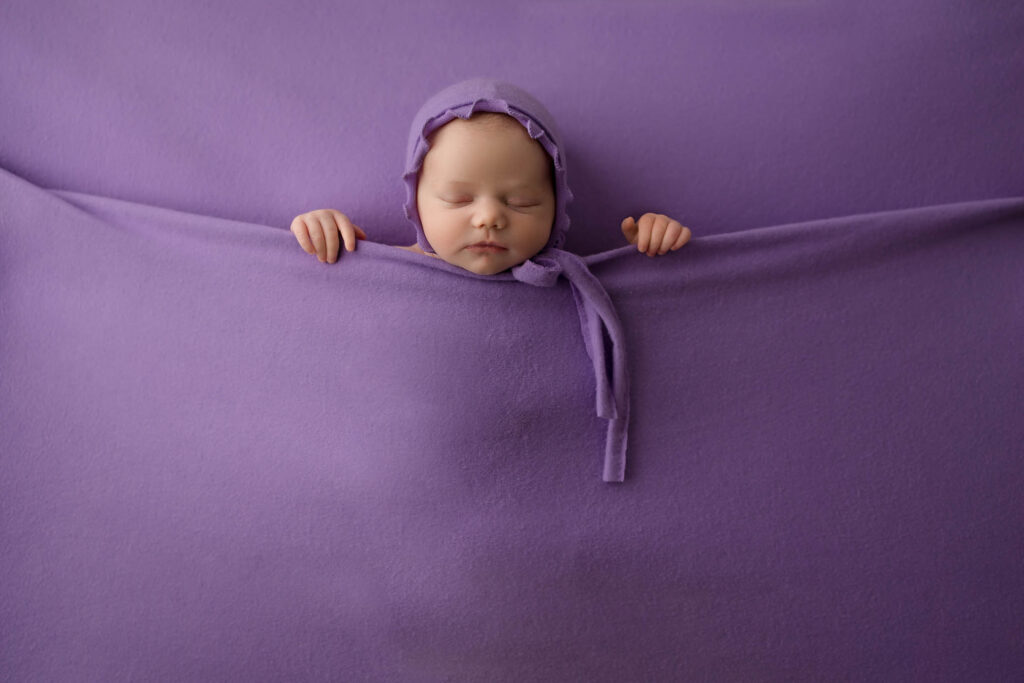 A sleeping baby wearing a purple hat is tucked snugly under a soft purple blanket, with only the head and hands visible, blending into the purple background—an enchanting moment captured by a skilled Phoenix newborn photographer.