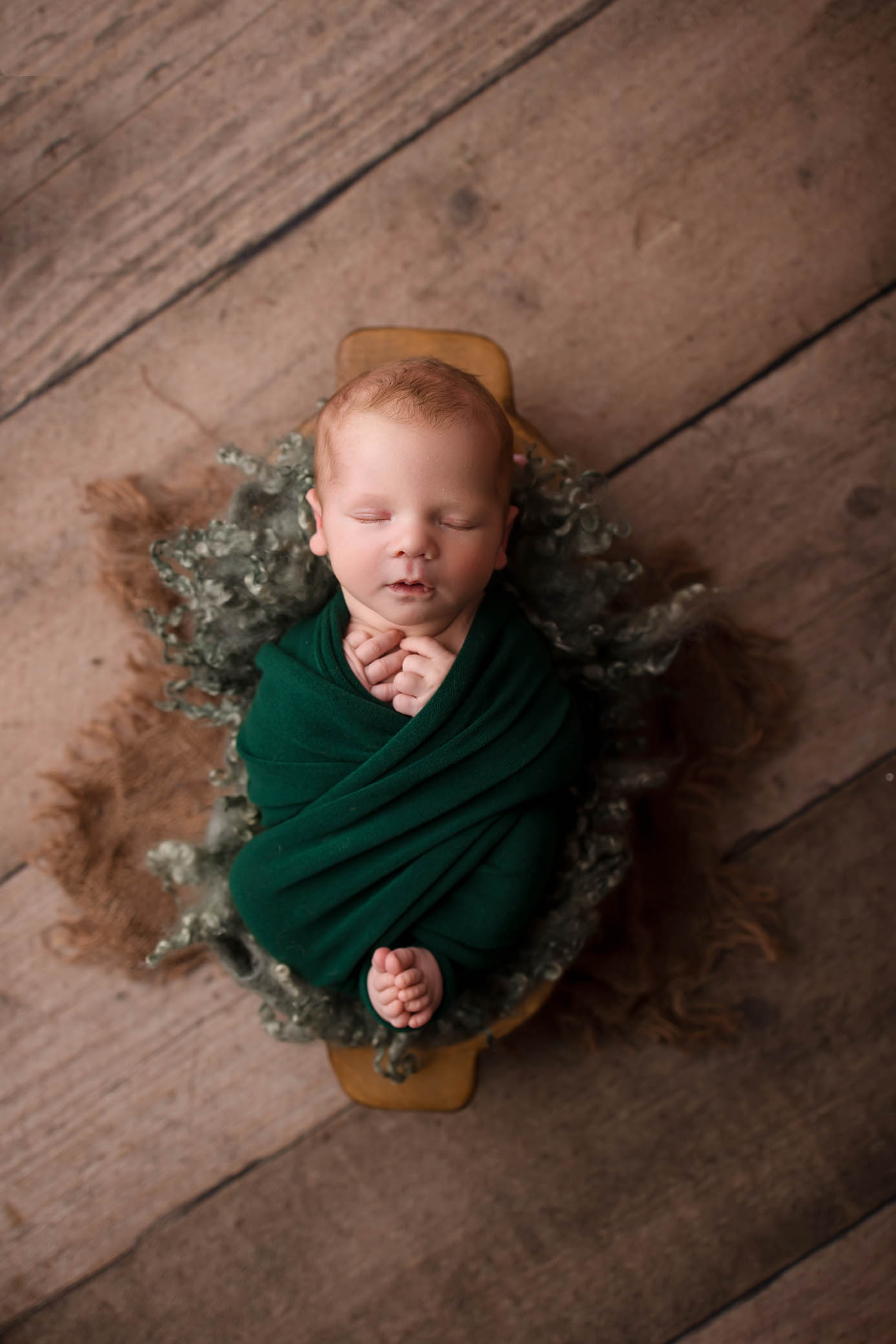 Newborn baby in dark green blanket on textured brown surface, eyes closed, hands and feet exposed.