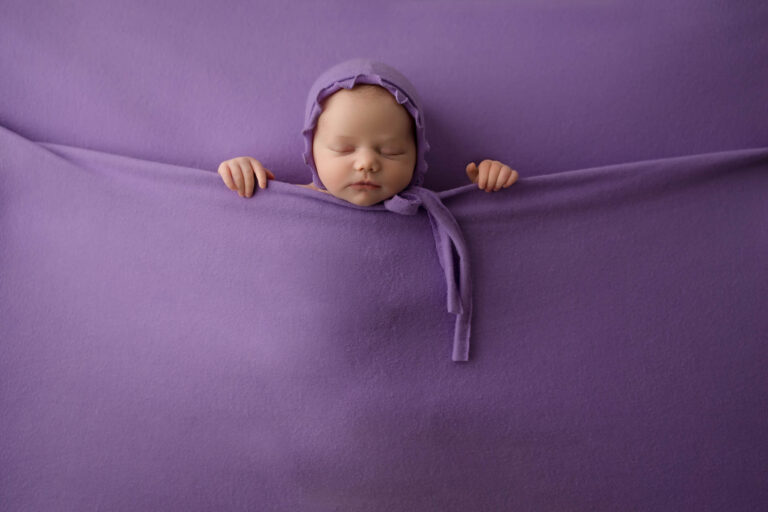 A sleeping baby wearing a purple hat is tucked snugly under a soft purple blanket, with only the head and hands visible, blending into the purple background—an enchanting moment captured by a skilled Phoenix newborn photographer.