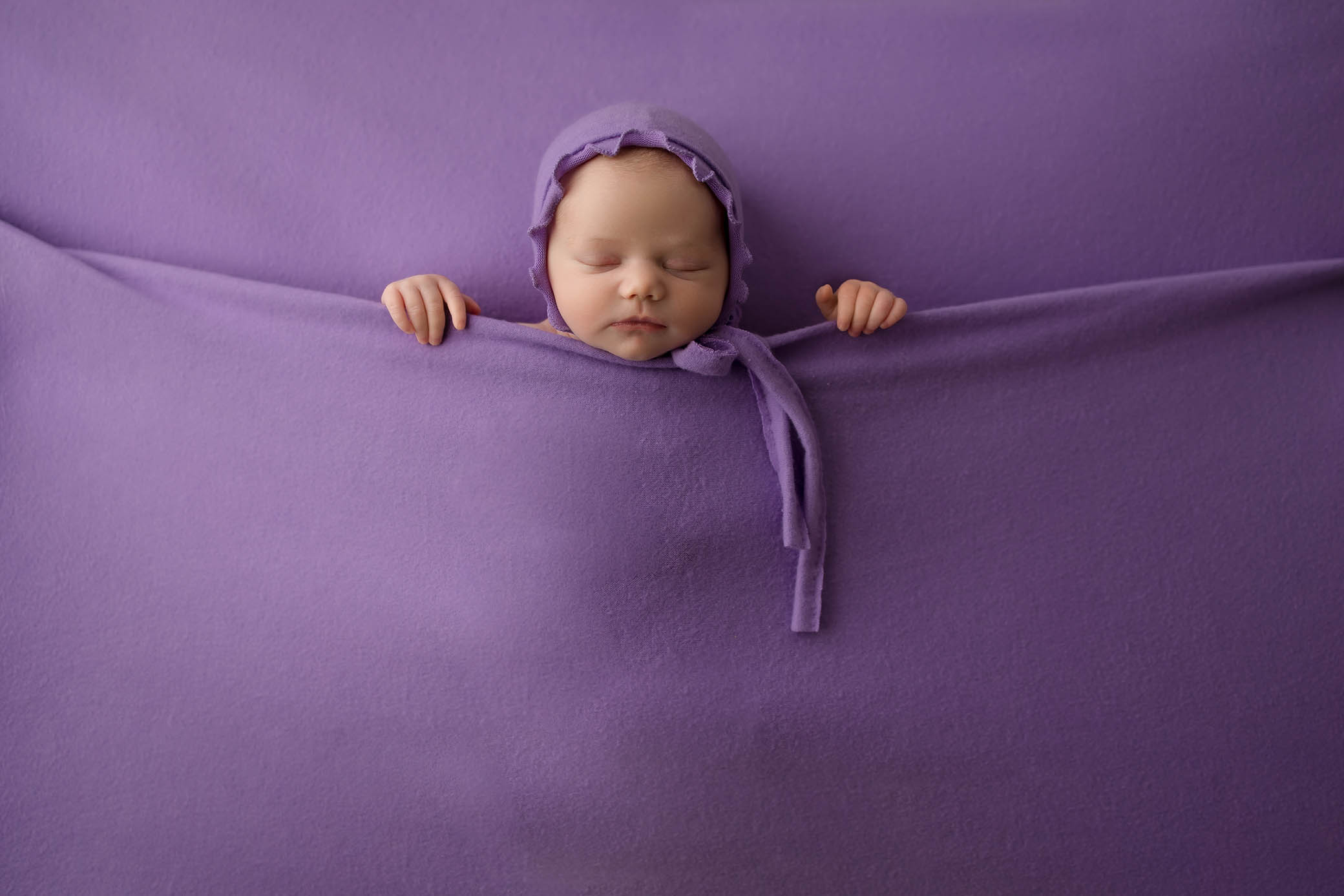 A sleeping baby wearing a purple hat is tucked snugly under a soft purple blanket, with only the head and hands visible, blending into the purple background—an enchanting moment captured by a skilled Phoenix newborn photographer.