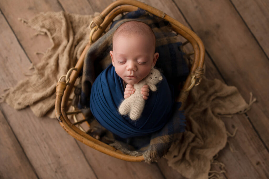 A newborn baby wrapped in a dark blue blanket sleeps peacefully in a wicker basket, holding a small knitted teddy bear—captured beautifully by a Phoenix newborn photographer—with a textured brown cloth beneath on a wooden floor.