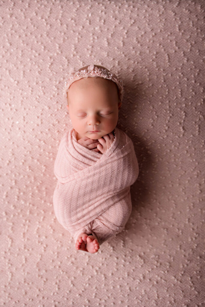 Newborn baby wrapped in a soft pink blanket, sleeping peacefully on a textured pink surface, with a delicate pink headband—captured beautifully by a skilled Phoenix newborn photographer.