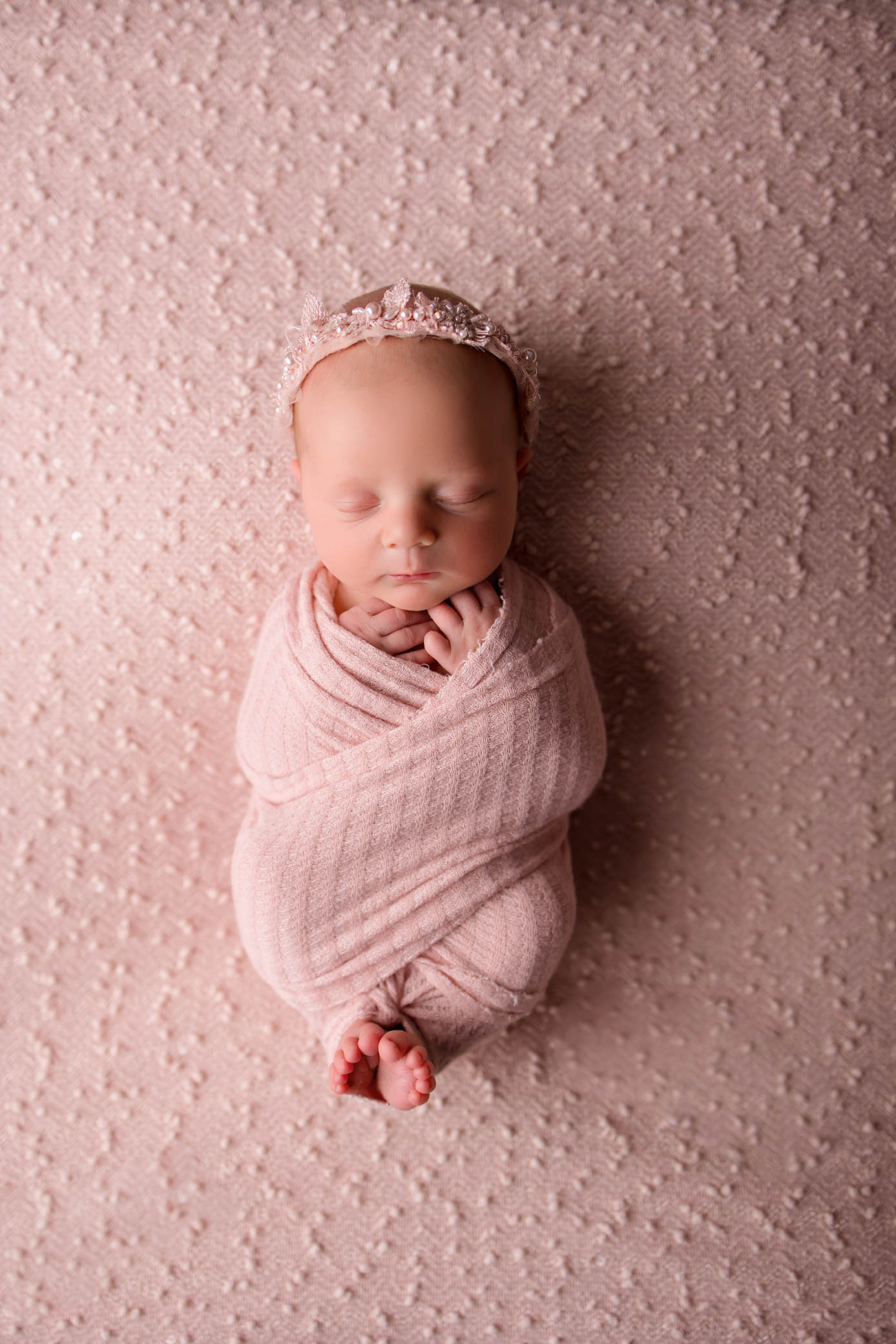 Newborn baby wrapped in a soft pink blanket, sleeping peacefully on a textured pink surface, with a delicate pink headband—captured beautifully by a skilled Phoenix newborn photographer.