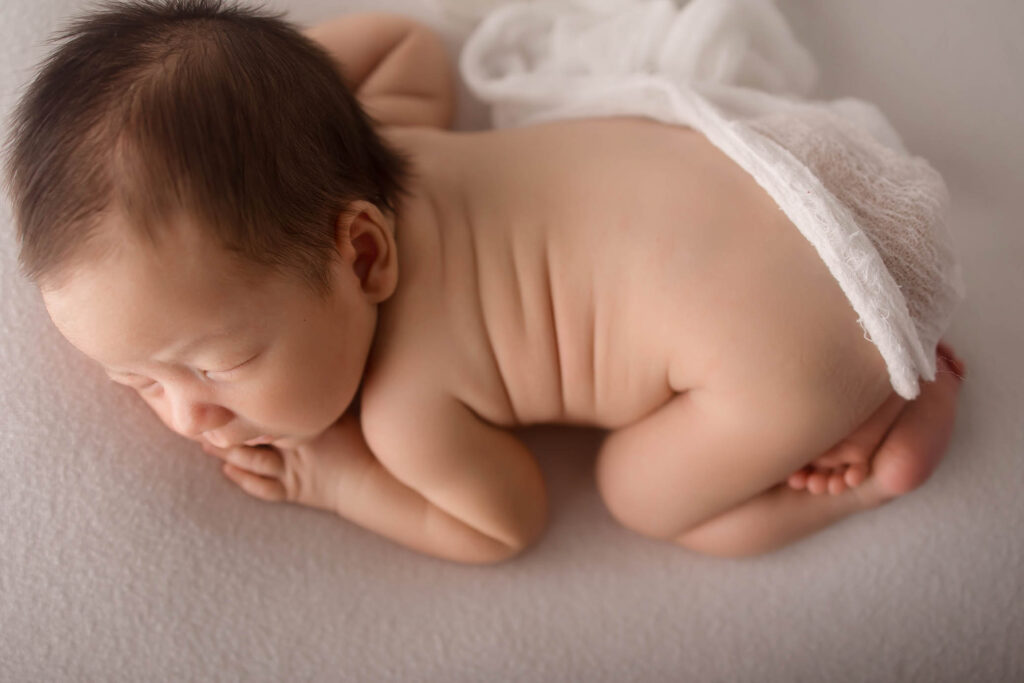 A phoenix newborn photographer captures a newborn baby lying on their stomach on a soft white blanket, with a sheer white cloth partially covering their lower back and legs. The baby is sleeping peacefully with arms folded under their head.