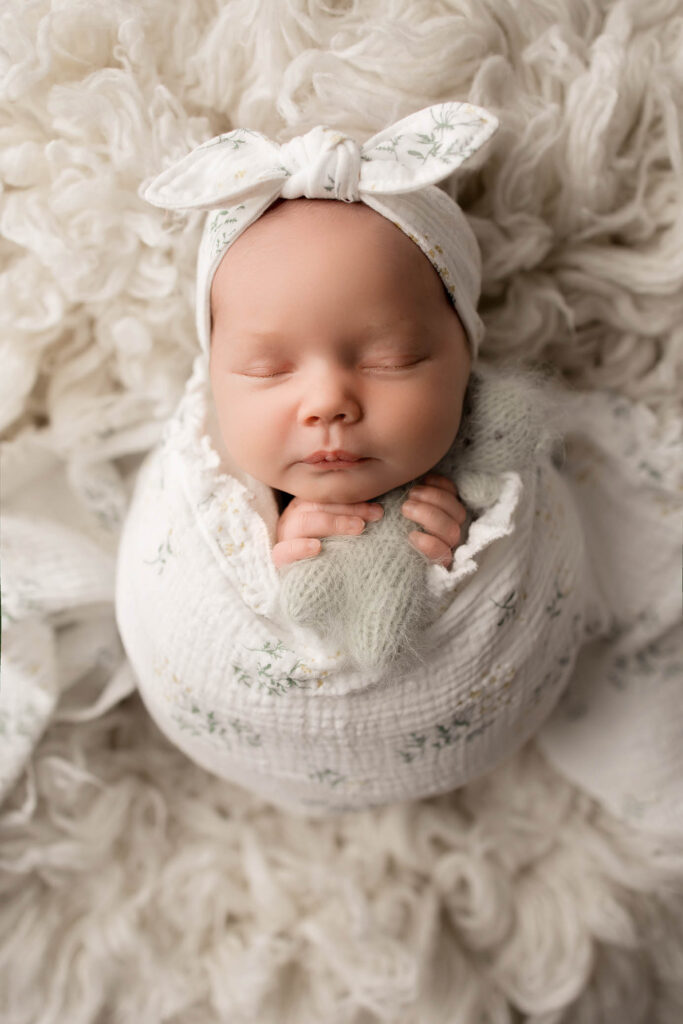 A newborn baby wrapped in a white blanket with delicate patterns, wearing a matching headband with a bow, sleeps peacefully on a fluffy white surface—captured by a talented Glendale maternity photographer.