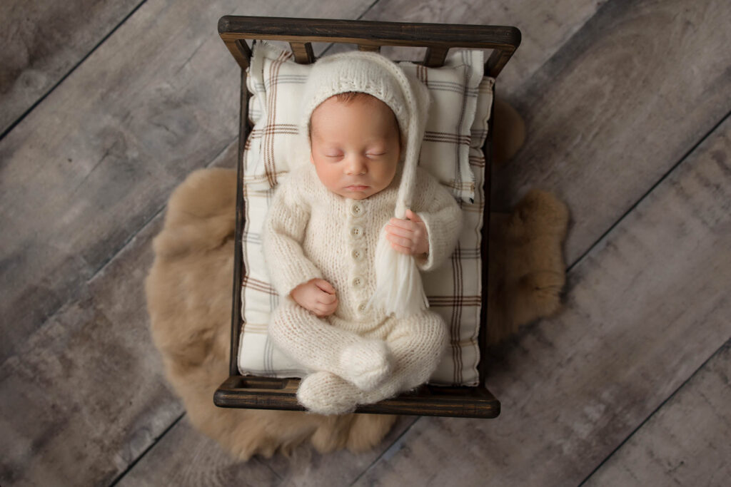 A newborn baby sleeps peacefully on a small, plaid cushion in a wooden bed, wearing a cozy cream knit outfit and matching hat—a heartwarming scene captured by a Peoria maternity photographer, with a soft brown rug beneath the bed on a wooden floor.