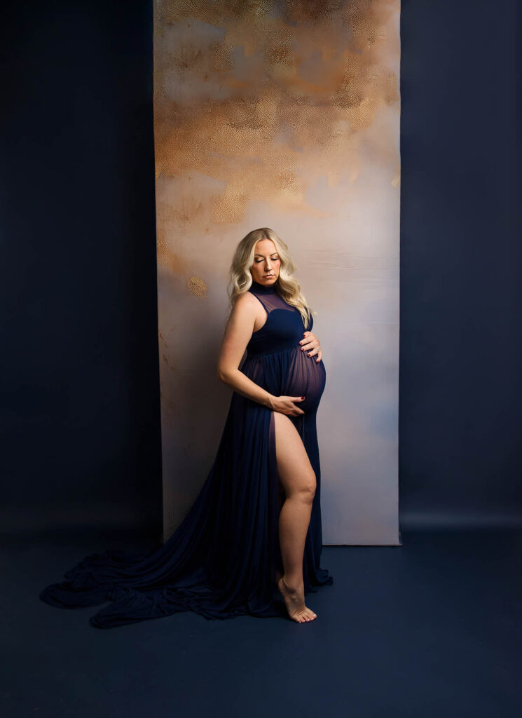 A pregnant woman in a flowing navy blue gown stands barefoot against a textured backdrop, gently holding her belly and looking down with a serene expression—captured by a talented Scottsdale maternity photographer.