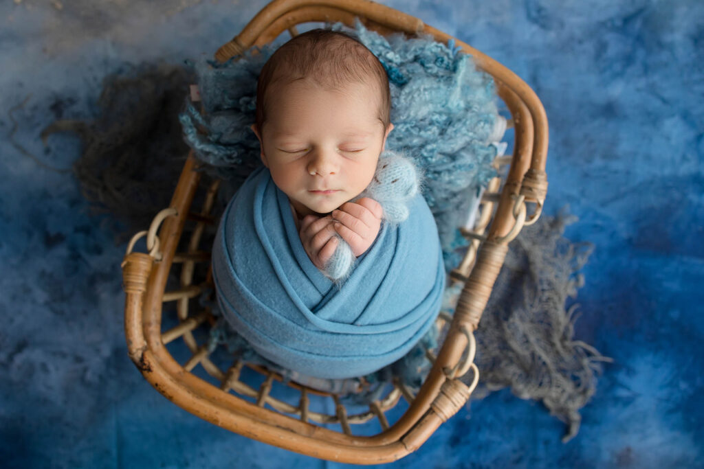 A newborn baby peacefully sleeps wrapped in a soft blue blanket, lying in a wicker basket with blue textured fabric and background—captured beautifully by a skilled Phoenix newborn photographer.