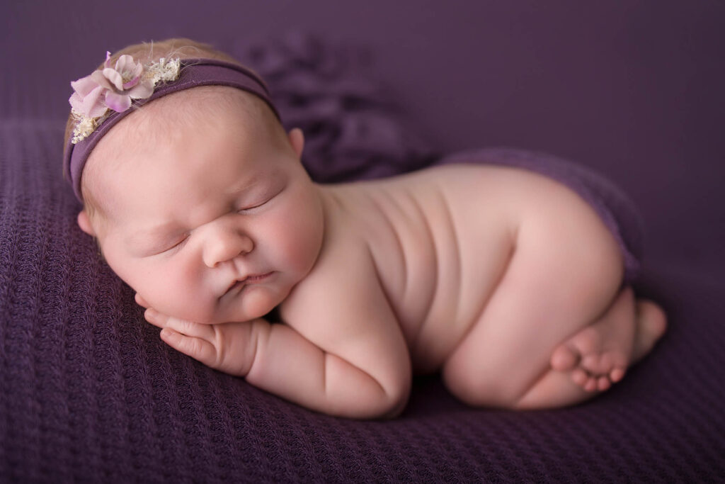 A sleeping baby wearing a purple headband with a flower lies curled up on a purple textured blanket, peacefully resting with eyes closed—captured beautifully by a Scottsdale maternity photographer.