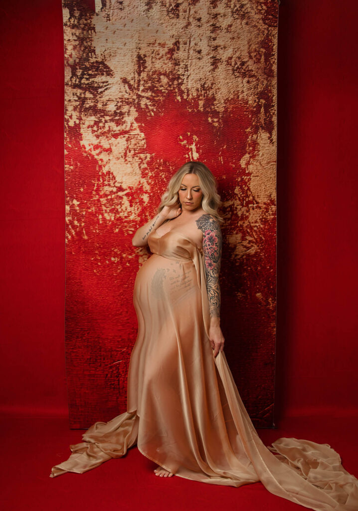 A pregnant woman with long blonde hair and a colorful tattoo on her arm stands barefoot on a red floor, wearing a sheer nude gown, in front of a textured red and gold backdrop, captured by a talented Glendale maternity photographer.