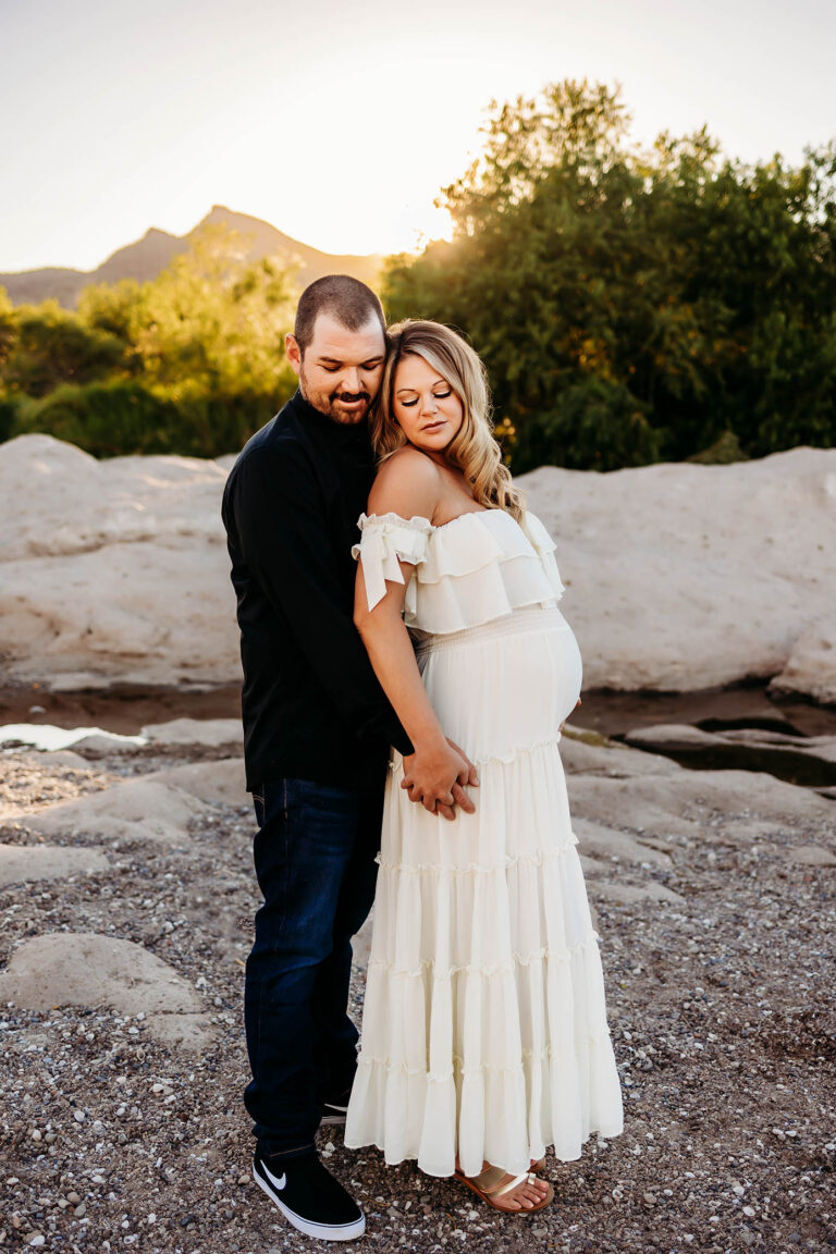 A couple stands outdoors on rocky ground at sunset. The man, in a black shirt and jeans, stands behind the pregnant woman in an off-shoulder white dress. As they gently hold hands over her baby bump, they embody the calm artistry of a phoenix maternity photographer.