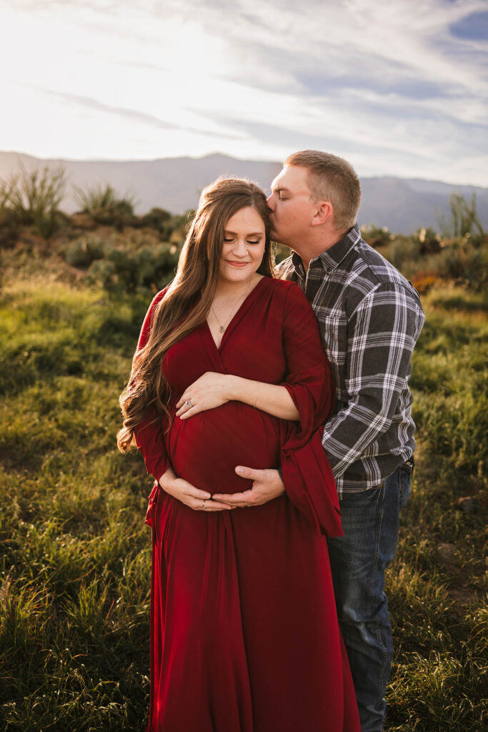 A man kisses a pregnant woman’s forehead while standing behind her in a sunlit field. The woman, in a long red dress, cradles her belly as they enjoy a peaceful moment—captured beautifully by a Glendale maternity photographer.
