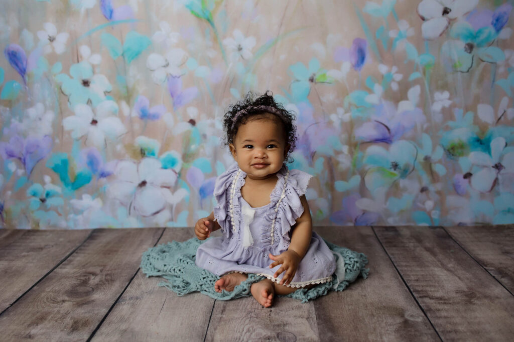 A smiling baby with curly hair sits on a teal blanket in a light purple dress. Pastel, floral-themed artwork decorates the background—captured beautifully by a talented Phoenix newborn photographer.
