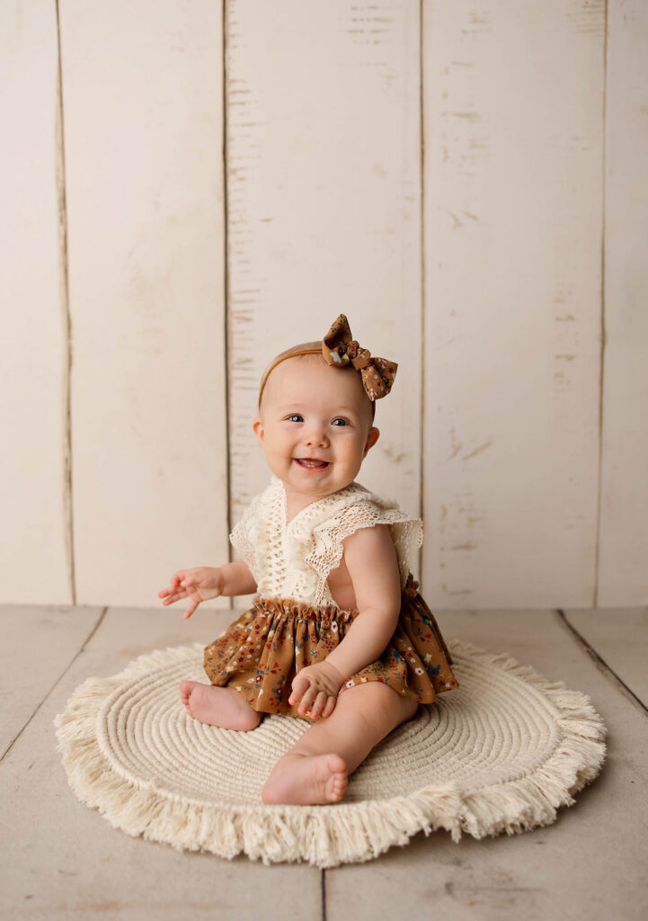 A smiling baby wearing a brown floral dress with a lace collar and matching bow sits on a round, fringed rug against a light wooden background, perfectly capturing the style of a Peoria maternity photographer.