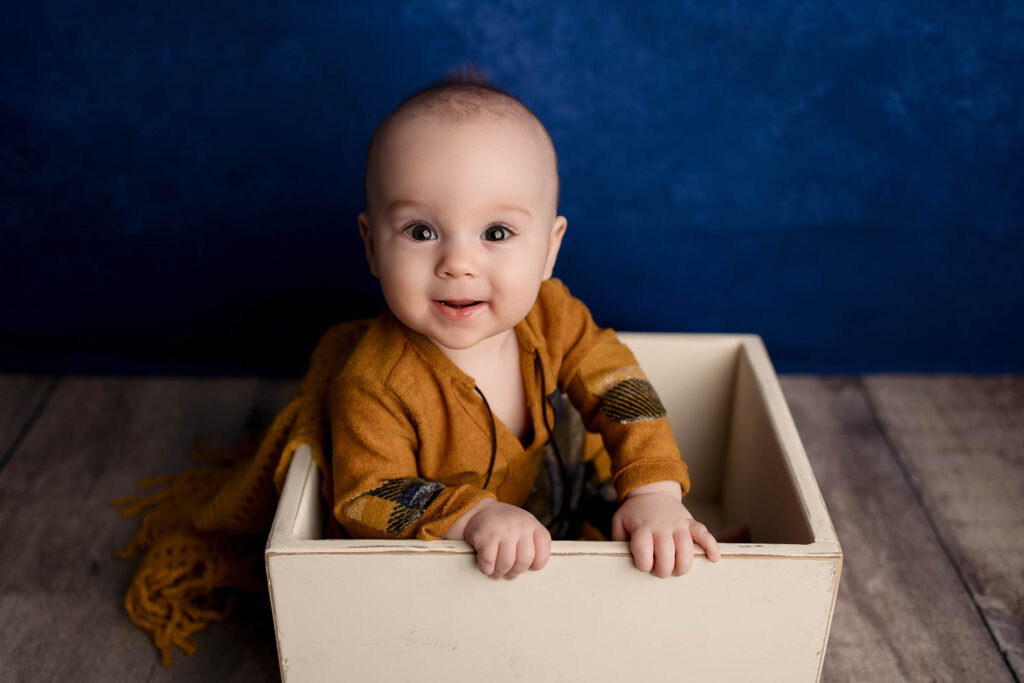 A smiling baby in a mustard yellow outfit sits inside a white wooden box on a wooden floor, with a deep blue background behind them—captured by a talented Scottsdale maternity photographer.