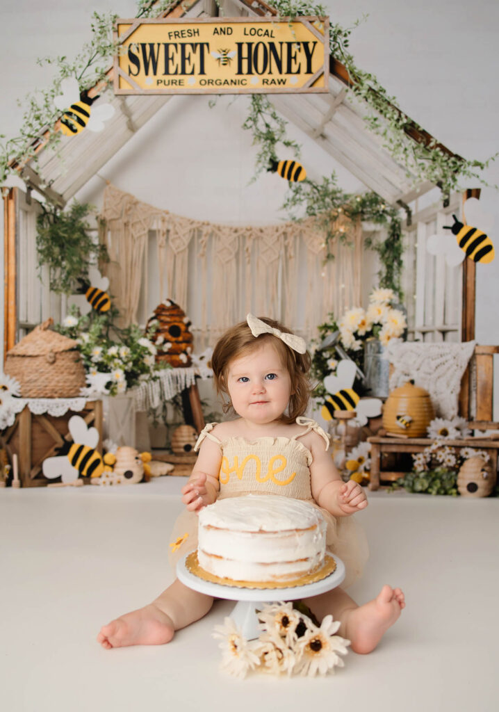 A toddler in a cream dress with "one" on it sits in front of a cake, surrounded by bee decorations and flowers, captured beautifully by a Phoenix newborn photographer with a rustic "Sweet Honey" sign and honey-themed backdrop behind her.