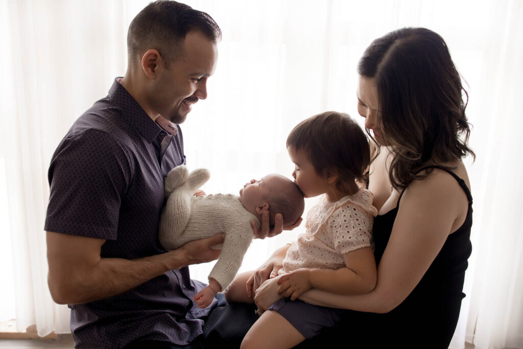 A man and woman sit close together, holding a toddler and a newborn baby. The toddler kisses the baby’s head as the adults smile warmly, bathed in soft window light—an intimate moment captured by a skilled family photographer Phoenix AZ.