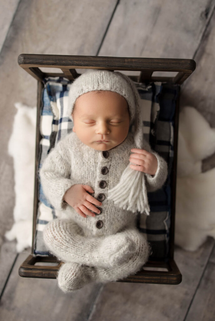 A newborn baby sleeps in a small wooden bed, wearing a cozy cream knit outfit and tassel hat. Captured by a Peoria maternity photographer, the baby rests on a blue and white plaid blanket on a wooden floor.