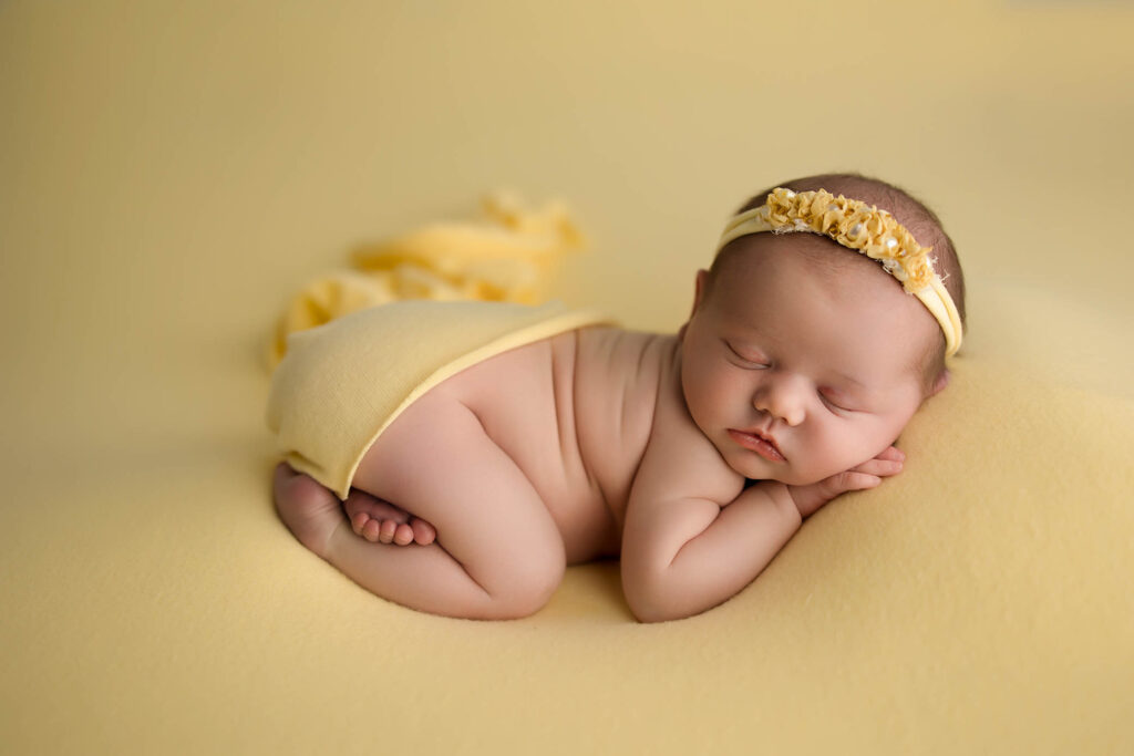 A newborn baby sleeps peacefully on a soft yellow blanket, wrapped in a yellow cloth with a flower headband—captured beautifully by a family photographer Phoenix AZ. The entire scene glows in warm yellow hues.