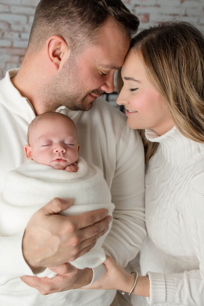 A man and woman wearing white sweaters lovingly cradle a sleeping swaddled newborn, their foreheads touching and smiling gently—an intimate moment captured by a family photographer in Phoenix, AZ.