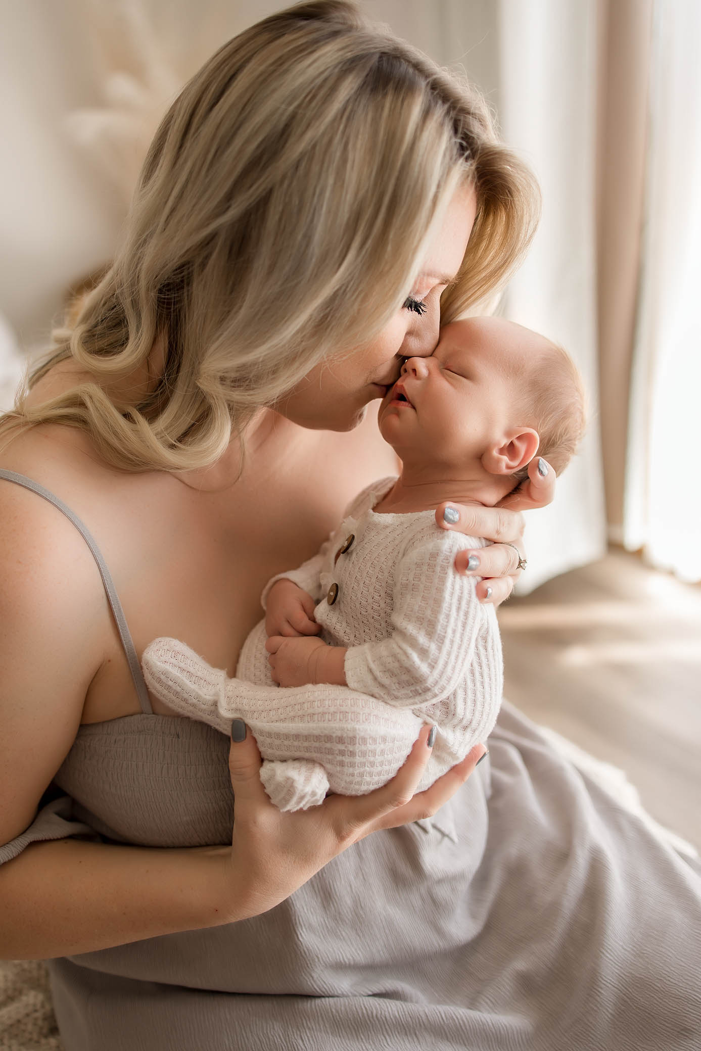 A woman in a light dress gently kisses the nose of a sleeping baby dressed in a cream knit outfit, holding the baby close in her arms in a softly lit, cozy room—captured by a skilled phoenix newborn photographer.