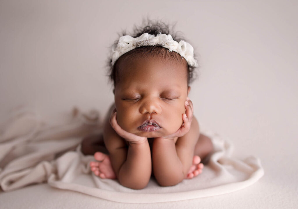 A newborn baby with closed eyes rests their head in their hands on a soft, cream-colored blanket. Captured by a Scottsdale maternity photographer, the baby wears a white floral headband and appears peaceful and relaxed.