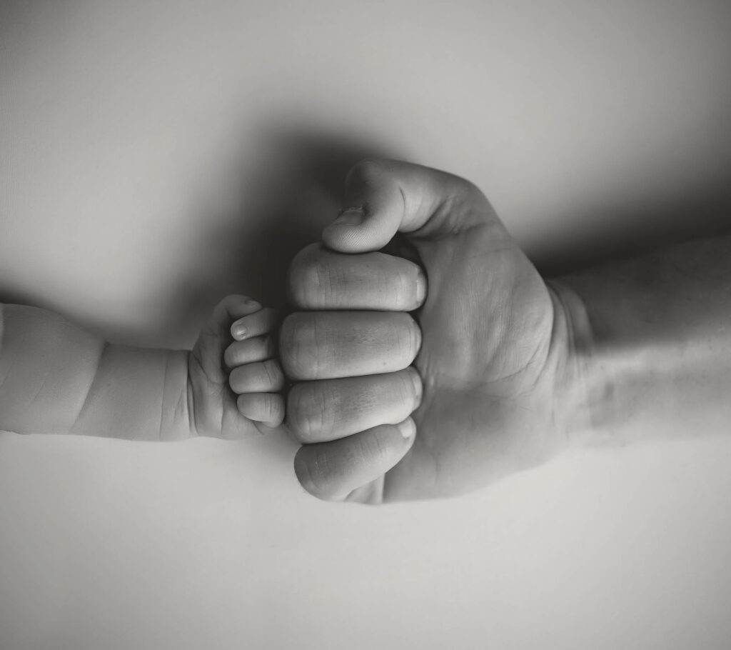 A black-and-white image shows a small child's hand gently holding the thumb of an adult hand, both forming fists and touching each other against a plain background—captured by a skilled family photographer in Phoenix, AZ.