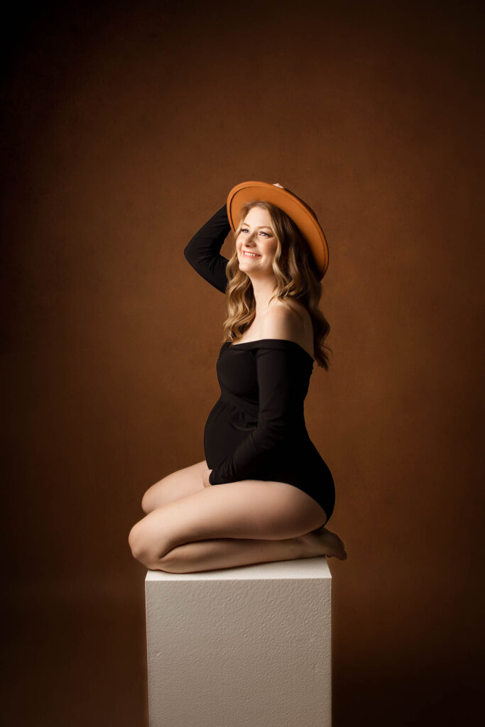 A smiling pregnant woman in a black off-shoulder bodysuit and orange hat kneels on a white cube against a brown studio background, captured beautifully by a Phoenix maternity photographer.
