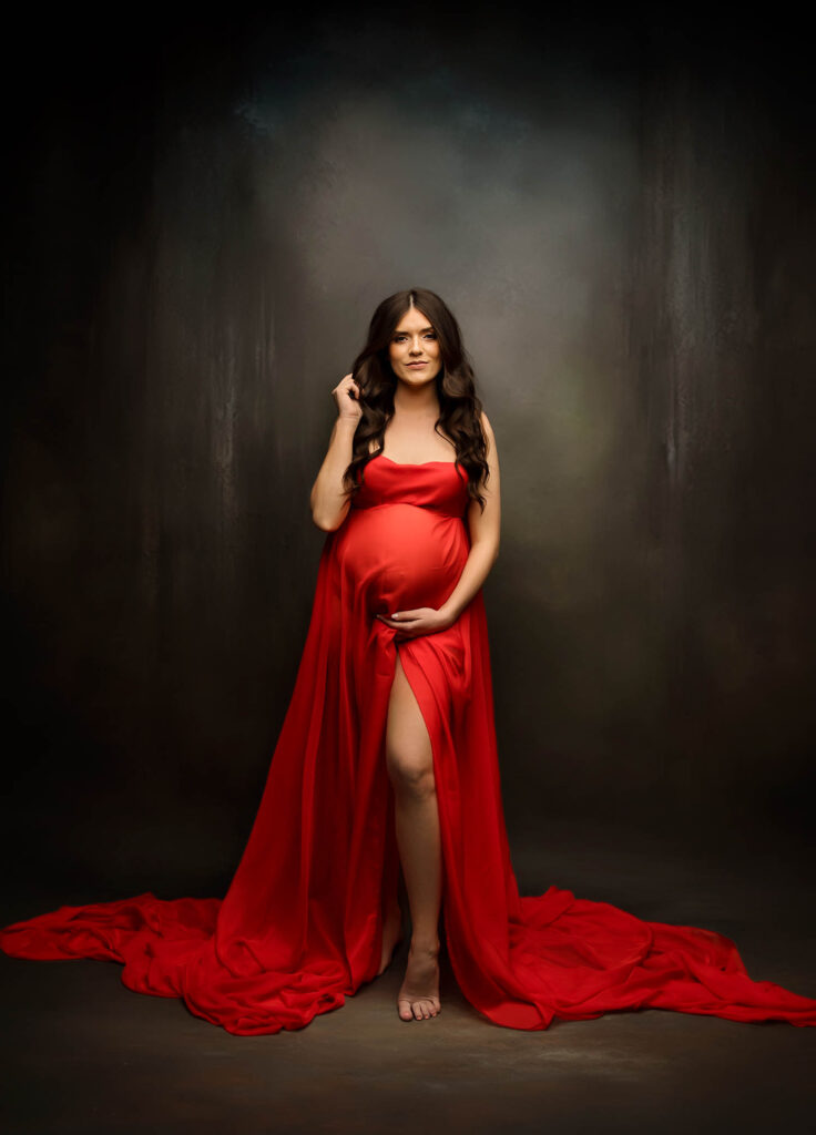 A pregnant woman with long dark hair stands barefoot in a flowing red gown, gently holding her belly, against a softly lit studio backdrop—captured beautifully by a skilled Scottsdale maternity photographer.