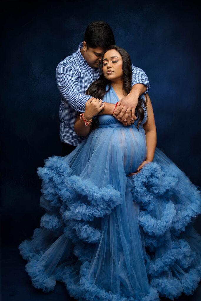 A couple poses in front of a dark blue background. The woman, visibly pregnant, wears a flowing blue tulle gown, while the man stands behind her, gently embracing her—a stunning moment captured by a Phoenix maternity photographer.